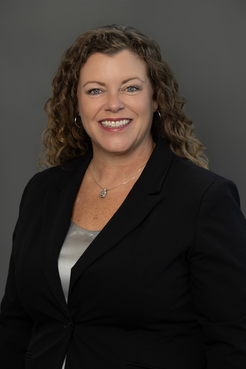 Smiling woman in a black blazer with a silver top, wearing a pendant necklace, against a gray backdrop (professional headshot).