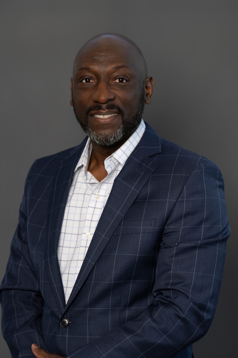 Head-and-shoulders portrait of a smiling Black man in a navy windowpane suit jacket and white grid shirt, neutral gray background.