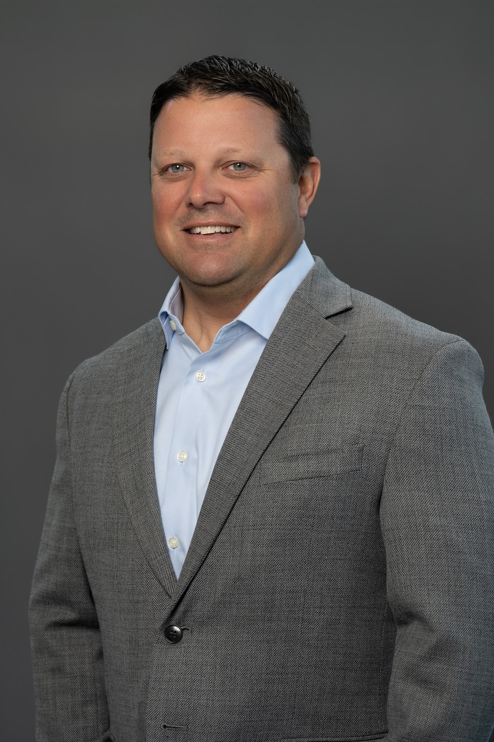 Professional headshot: smiling man in a gray blazer and light blue shirt against a neutral gray background.