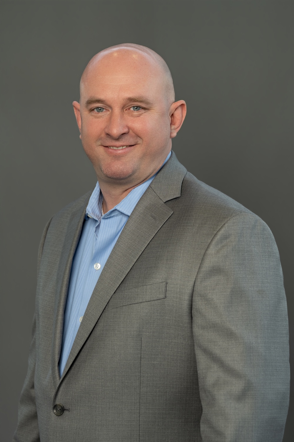 Head-and-shoulders portrait of a smiling bald man in a gray suit and light blue shirt, set against a plain gray backdrop.