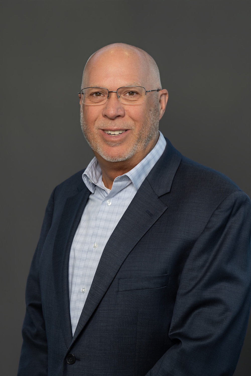 Smiling, bald man with glasses in a navy blazer and light shirt, posing for a headshot against a gray background.