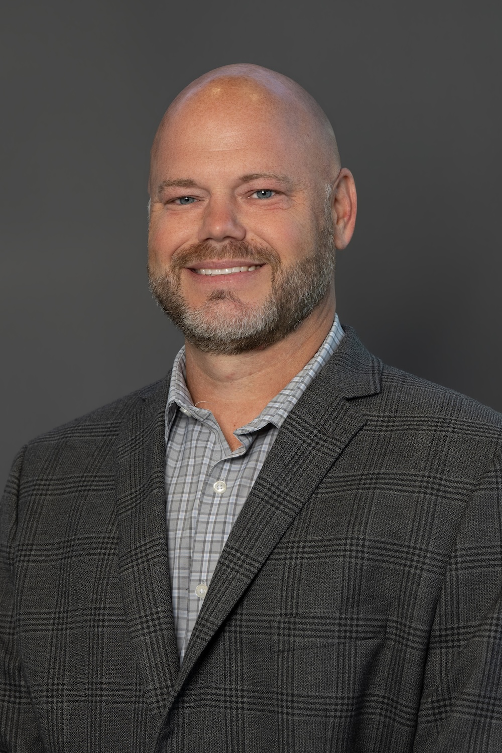 Professional head-and-shoulders portrait of a smiling bald man in a gray plaid blazer and light checkered shirt against a gray background.