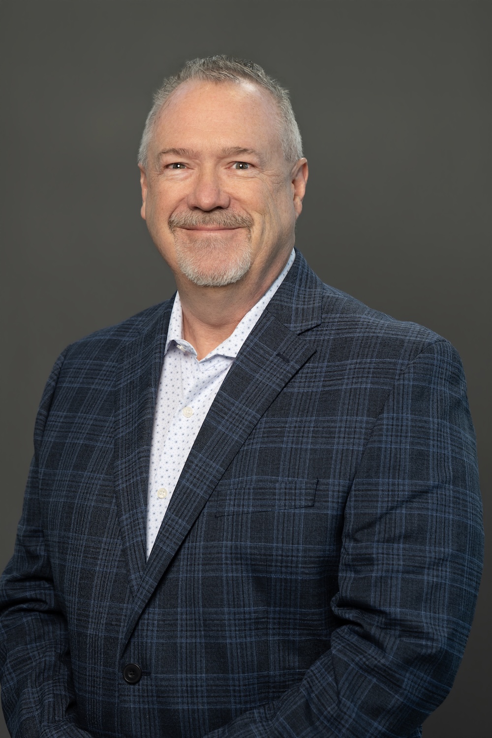 Portrait of a smiling middle-aged man with short gray hair and a goatee, wearing a blue plaid blazer and white dotted shirt against a neutral gray backdrop.