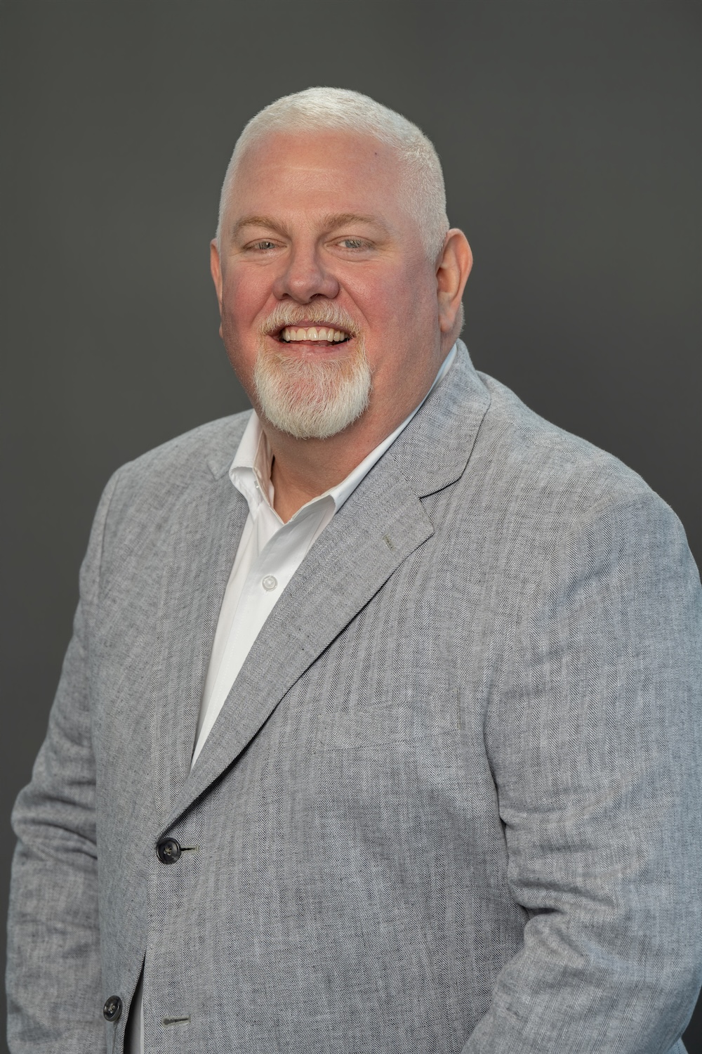 Smiling man in a light gray blazer and white shirt, head-and-shoulders portrait against a dark gray backdrop.