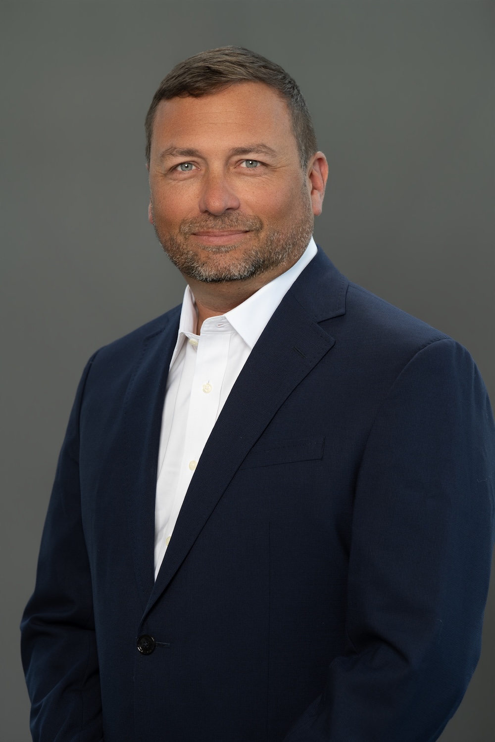 Professional headshot of a man in a navy blazer and white shirt against a gray studio backdrop, smiling slightly.