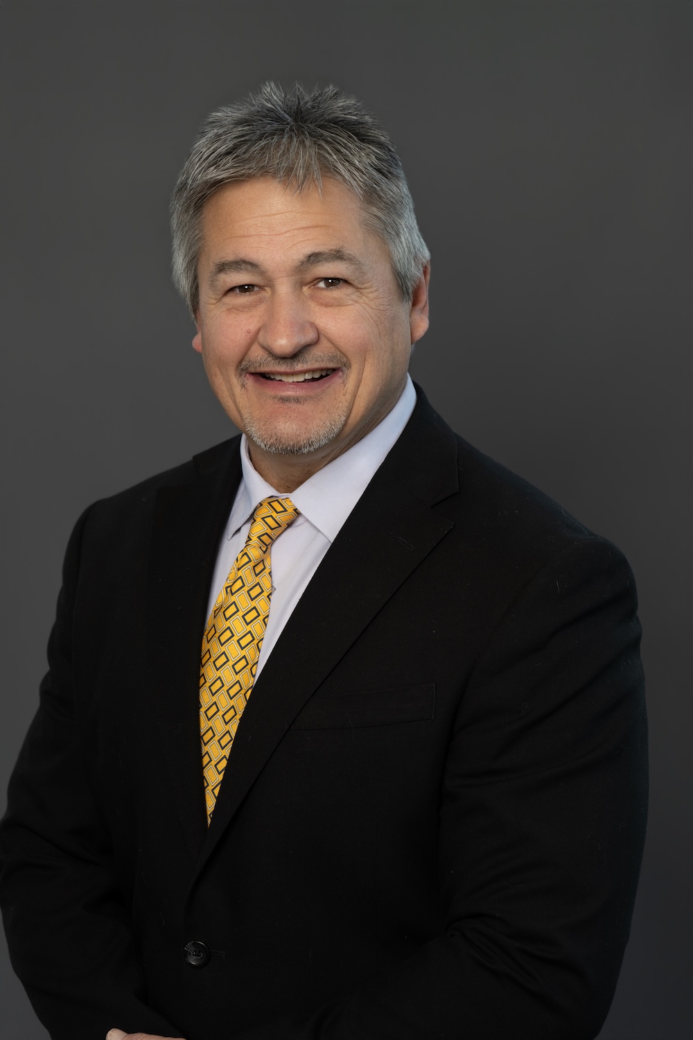 Professional headshot of a smiling man in a black suit and yellow patterned tie against a gray backdrop.