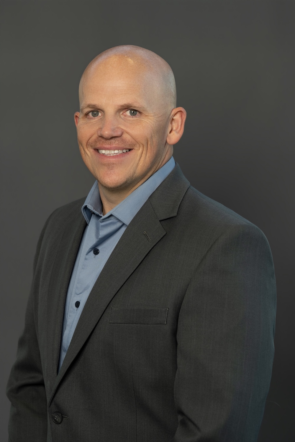 Smiling bald man in a dark suit and light blue shirt, posing for a professional headshot against a gray backdrop.