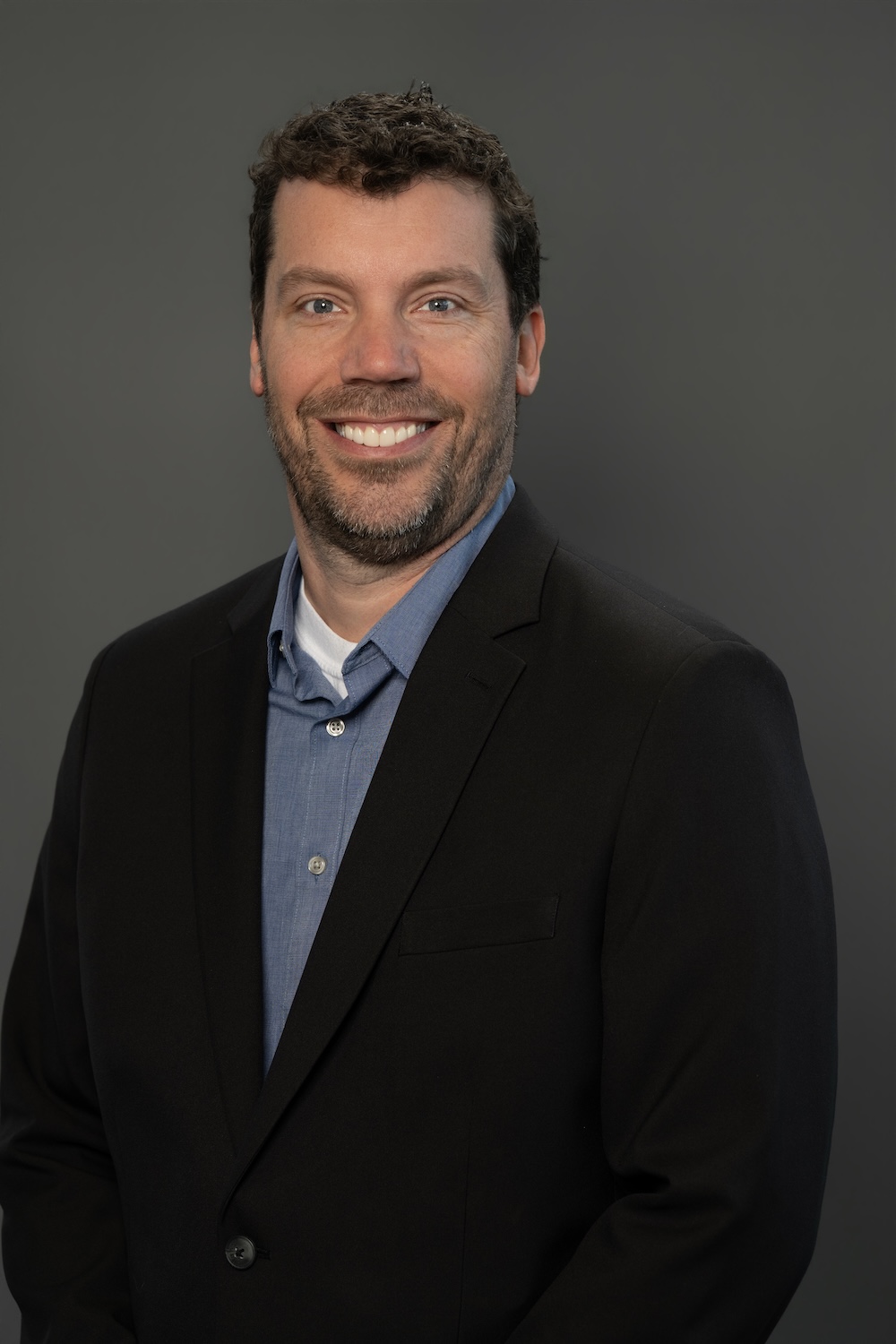Smiling man in a black blazer and blue shirt posing for a professional headshot against a gray backdrop.