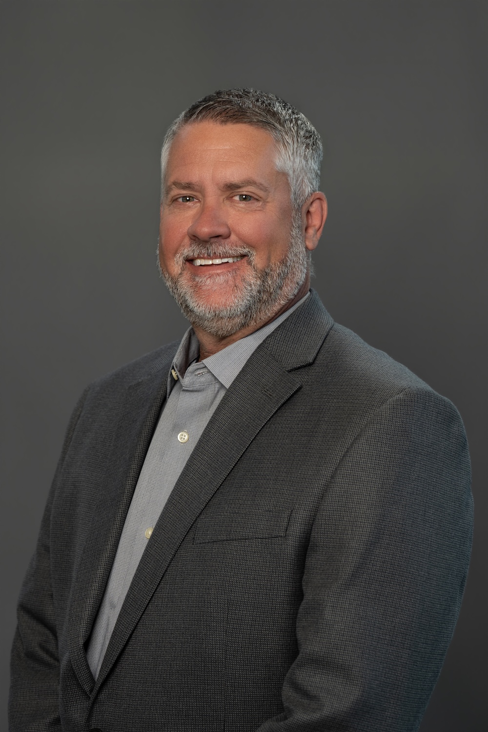Head-and-shoulders portrait of a smiling man in a gray blazer and light shirt against a dark gray background, a professional headshot.