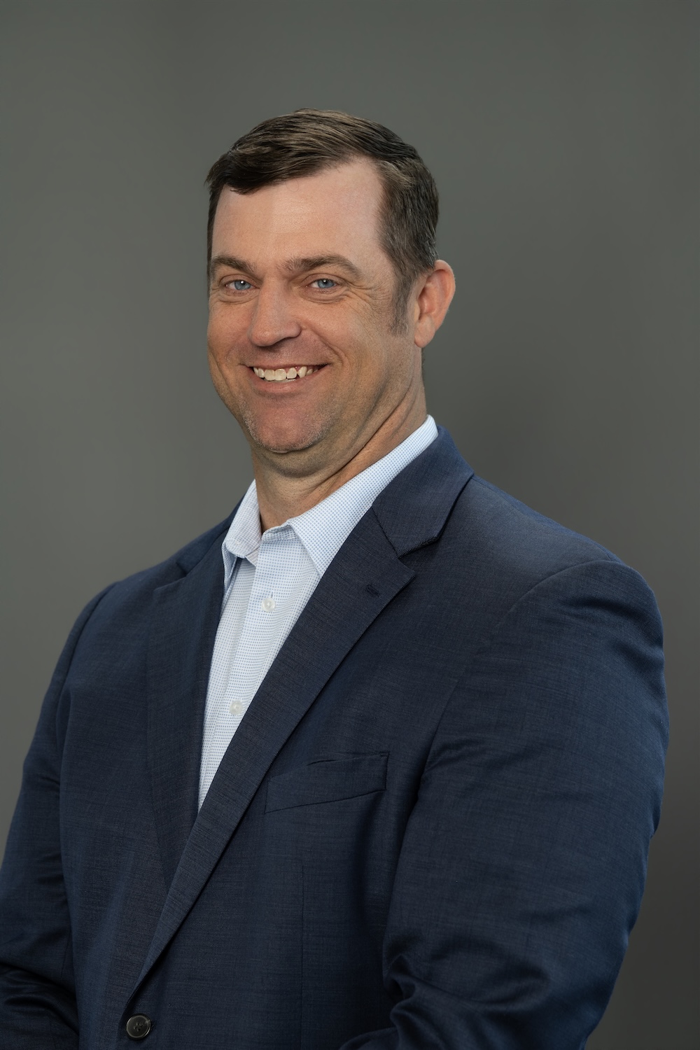 Smiling man in a navy blazer and light blue shirt poses for a professional headshot against a gray backdrop.