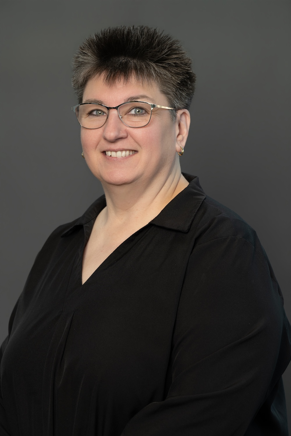 Smiling woman with short gray spiky hair and glasses in a black blouse against a gray backdrop.