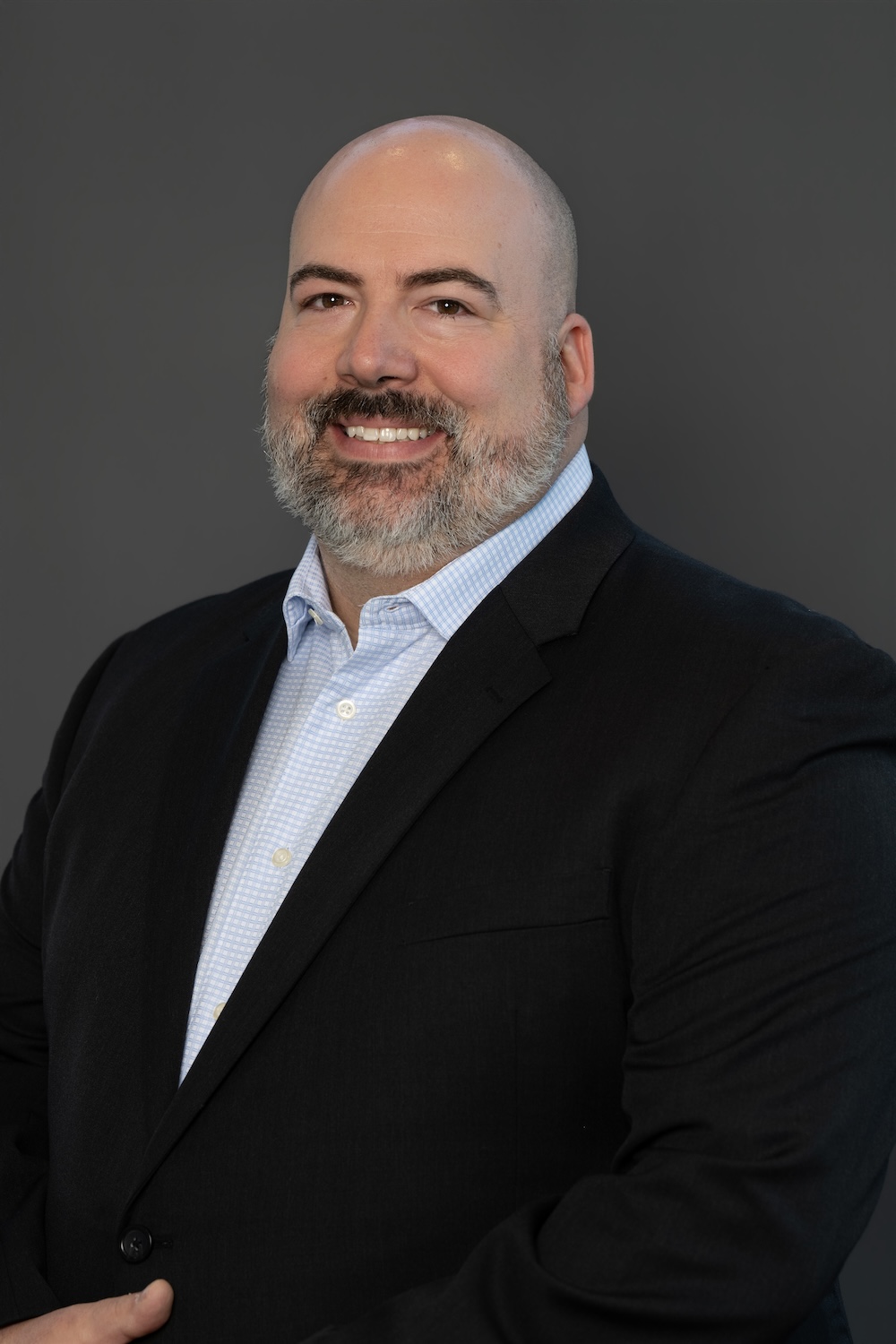 Smiling man in a black blazer and light blue checkered shirt against a gray studio background (professional headshot).