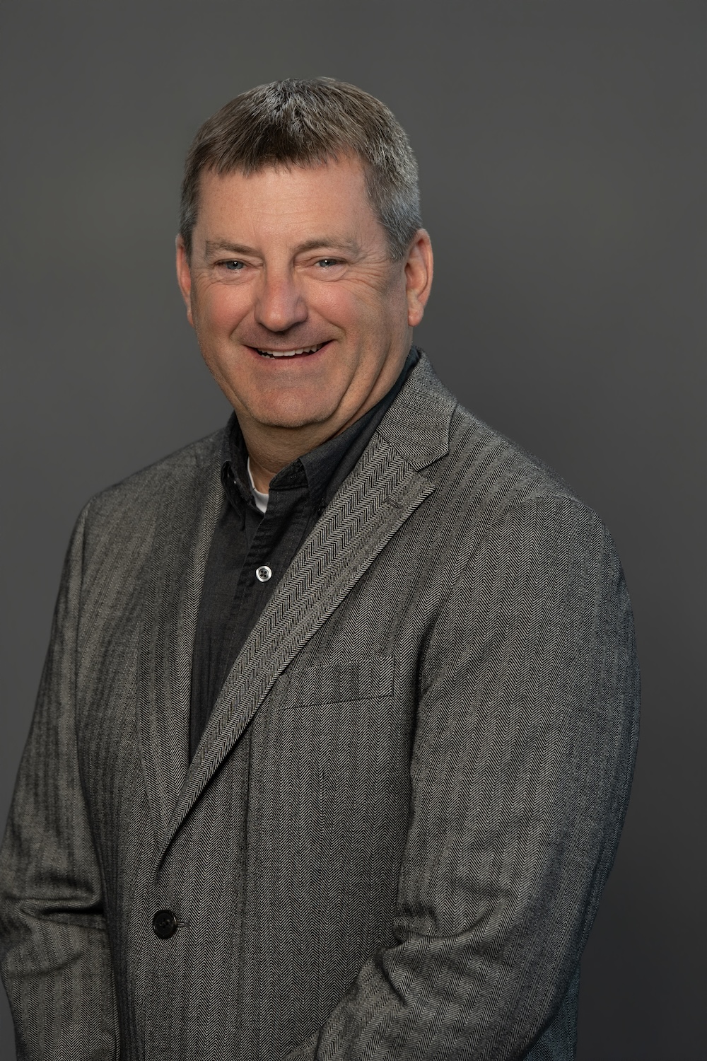 Head-and-shoulder portrait of a smiling man in a gray herringbone blazer against a dark gray background.