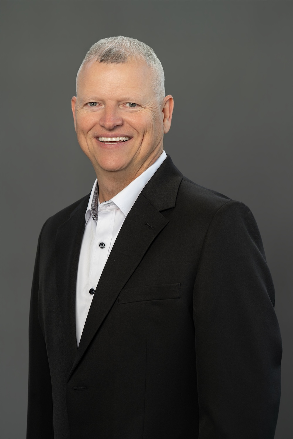 Smiling man in a black suit and white shirt poses for a professional headshot against a gray background.