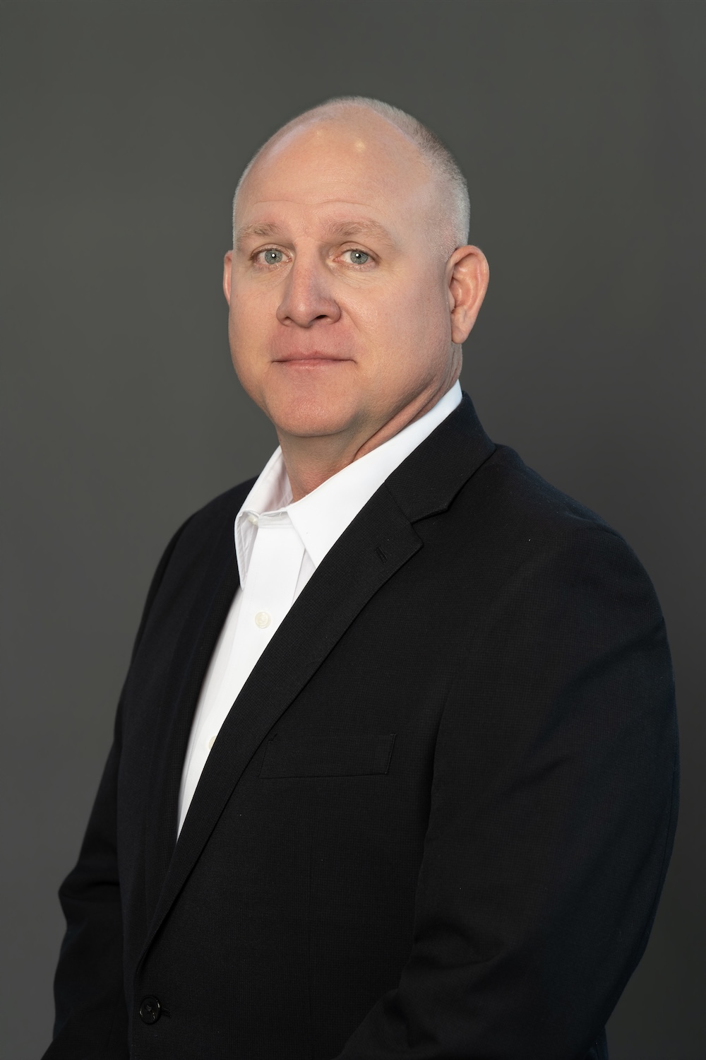 Professional headshot of a bald man in a black suit and white shirt, looking at the camera against a gray backdrop.