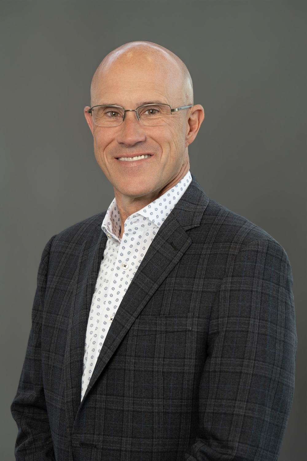 Professional headshot of a bald man wearing glasses and a dark plaid blazer, smiling at the camera.