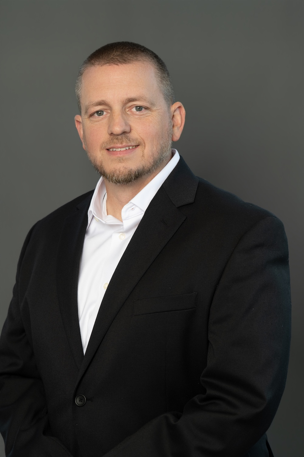 Professional head-and-shoulders portrait of a man in a black blazer and white shirt against a gray backdrop.