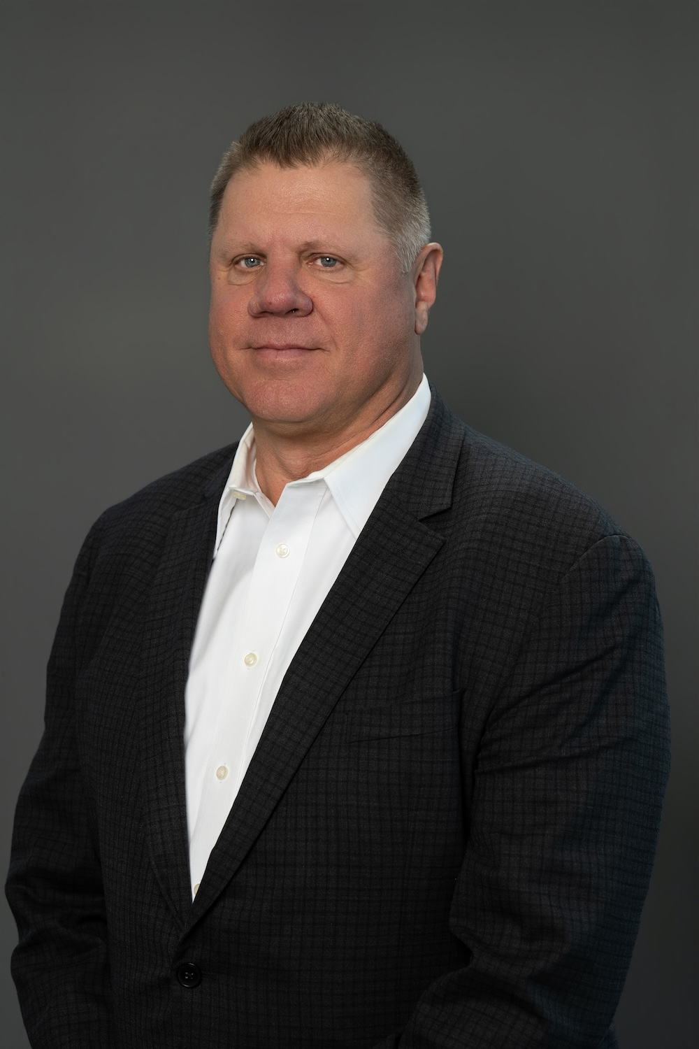Professional headshot of a middle-aged man in a dark blazer and white shirt, posed against a gray backdrop.