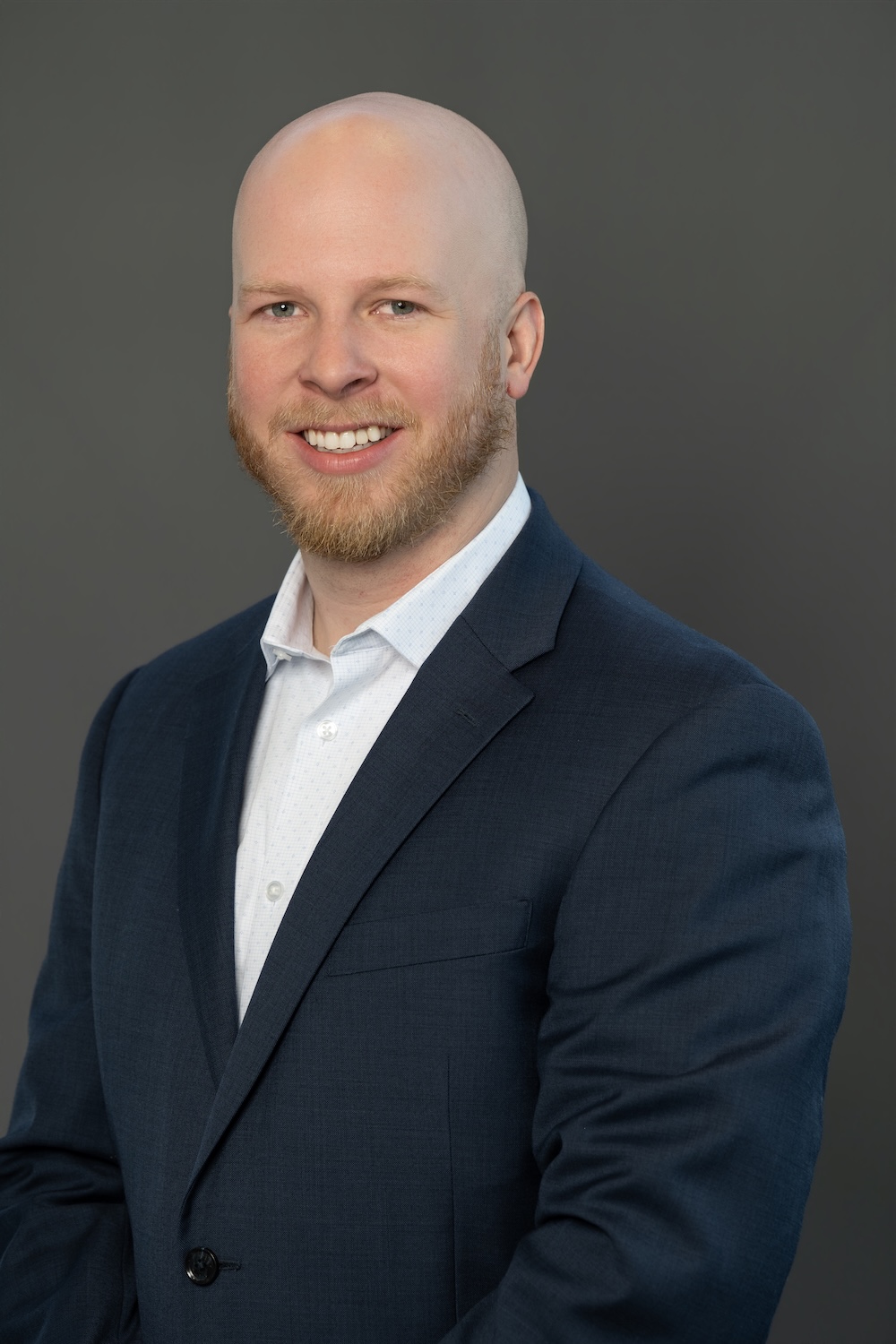 Head-and-shoulders portrait of a bald man with a red beard in a navy blazer, smiling against a gray background.
