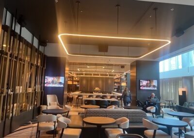 Modern lounge interior with a large glowing rectangular ring light, round tables and assorted seating, glass partitions on the left and windows on the right.