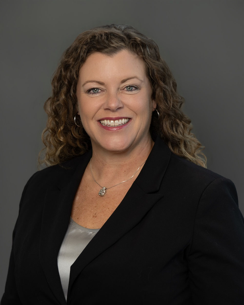 Angie-Kath-edit-2 Smiling woman in a black blazer with a silver top, wearing a pendant necklace, against a gray backdrop (professional headshot).