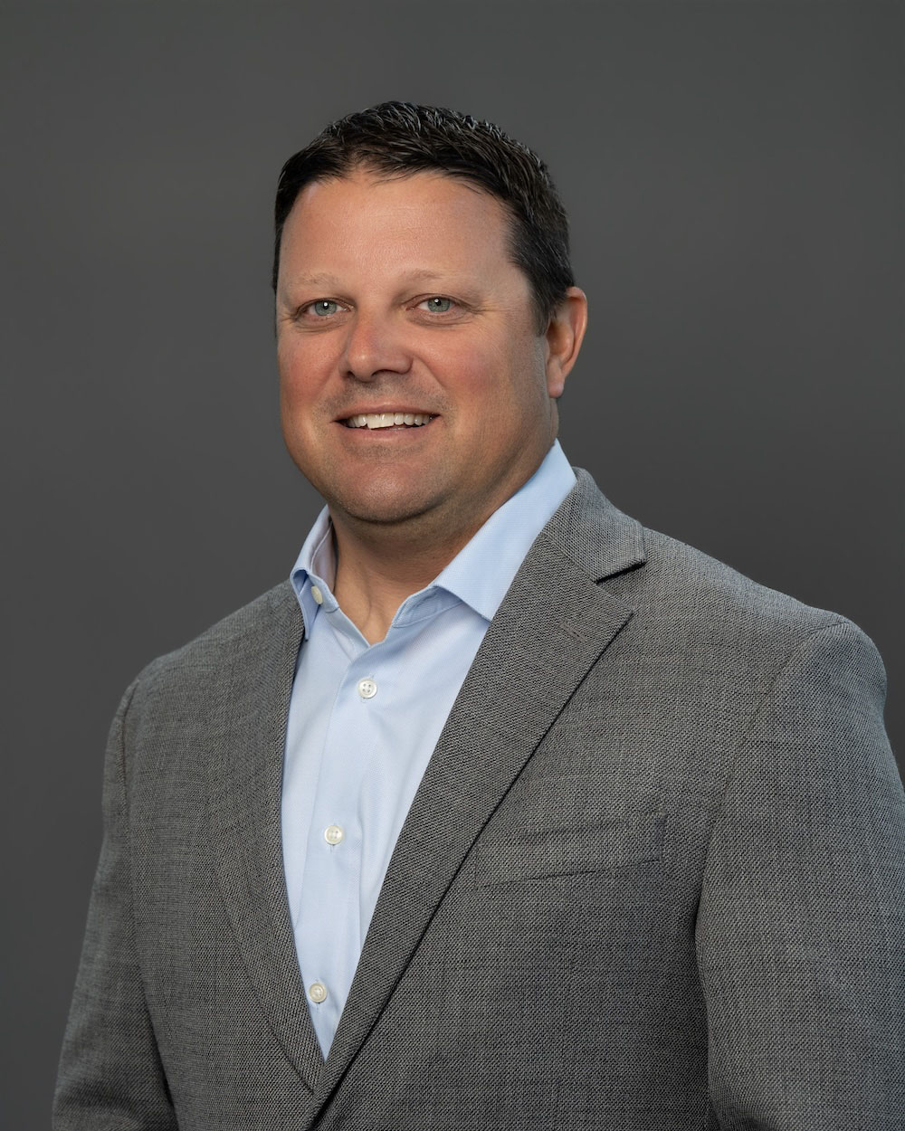 Austin-Schultz-edit-2 Professional headshot: smiling man in a gray blazer and light blue shirt against a neutral gray background.