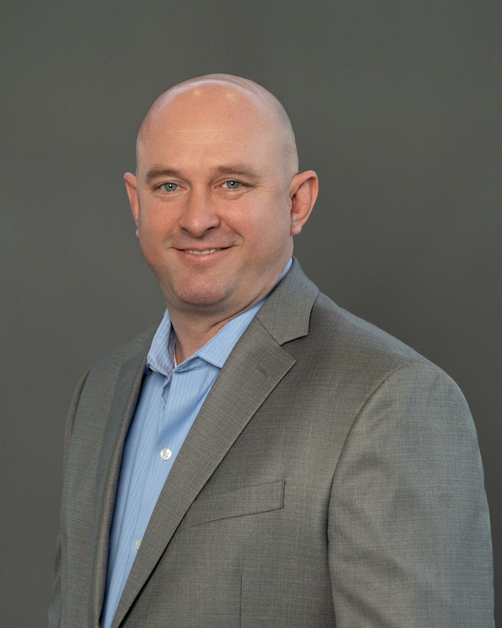 Brad-Niemantsverdriet-edit-2 Head-and-shoulders portrait of a smiling bald man in a gray suit and light blue shirt, set against a plain gray backdrop.