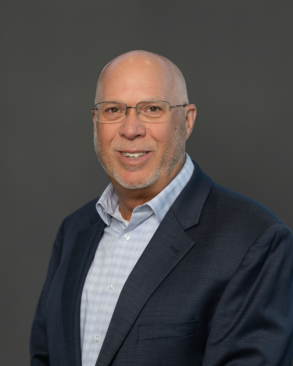 David-Nelson-edit-2 Smiling, bald man with glasses in a navy blazer and light shirt, posing for a headshot against a gray background.