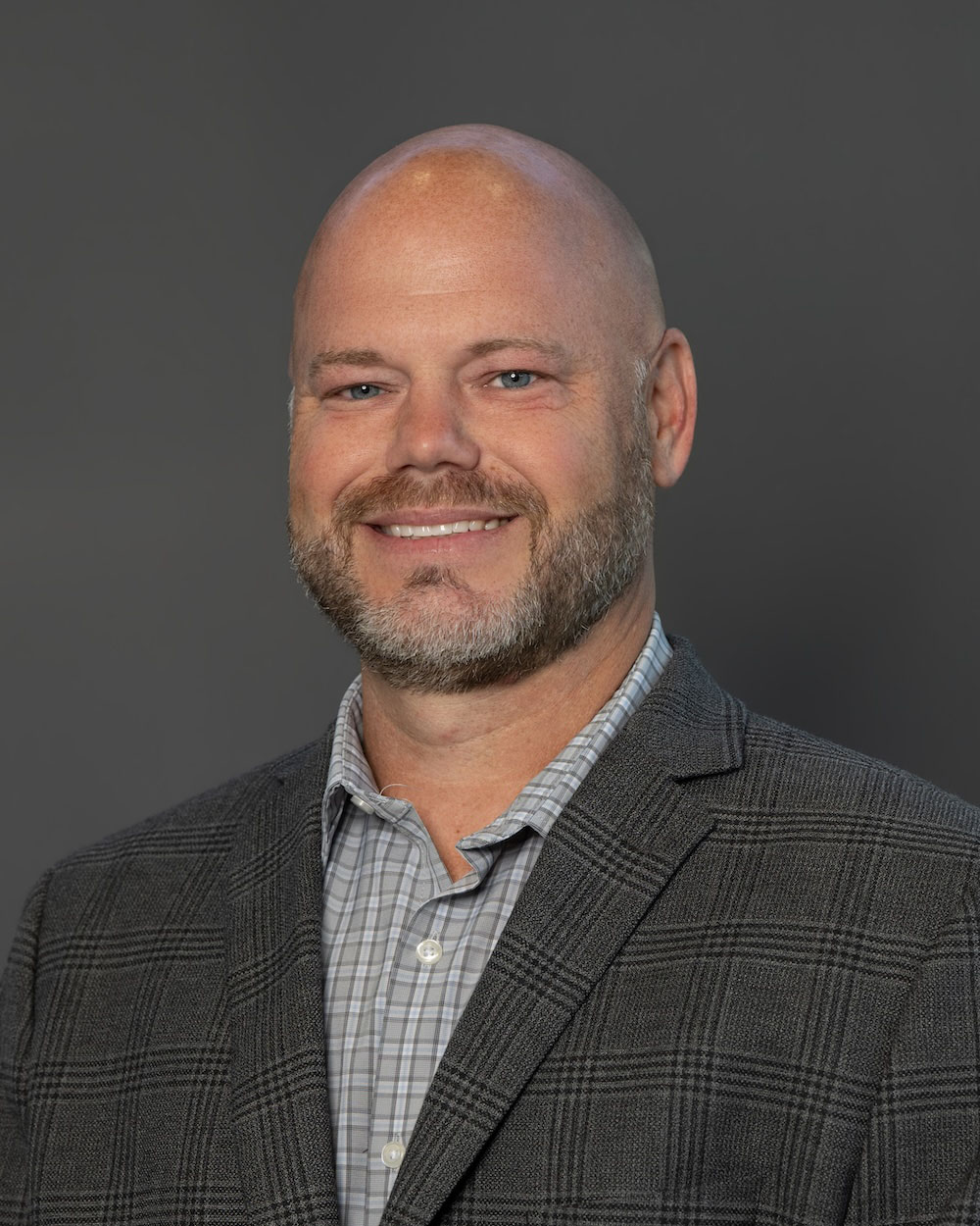 Frank-Gillett-edit-2 Professional head-and-shoulders portrait of a smiling bald man in a gray plaid blazer and light checkered shirt against a gray background.