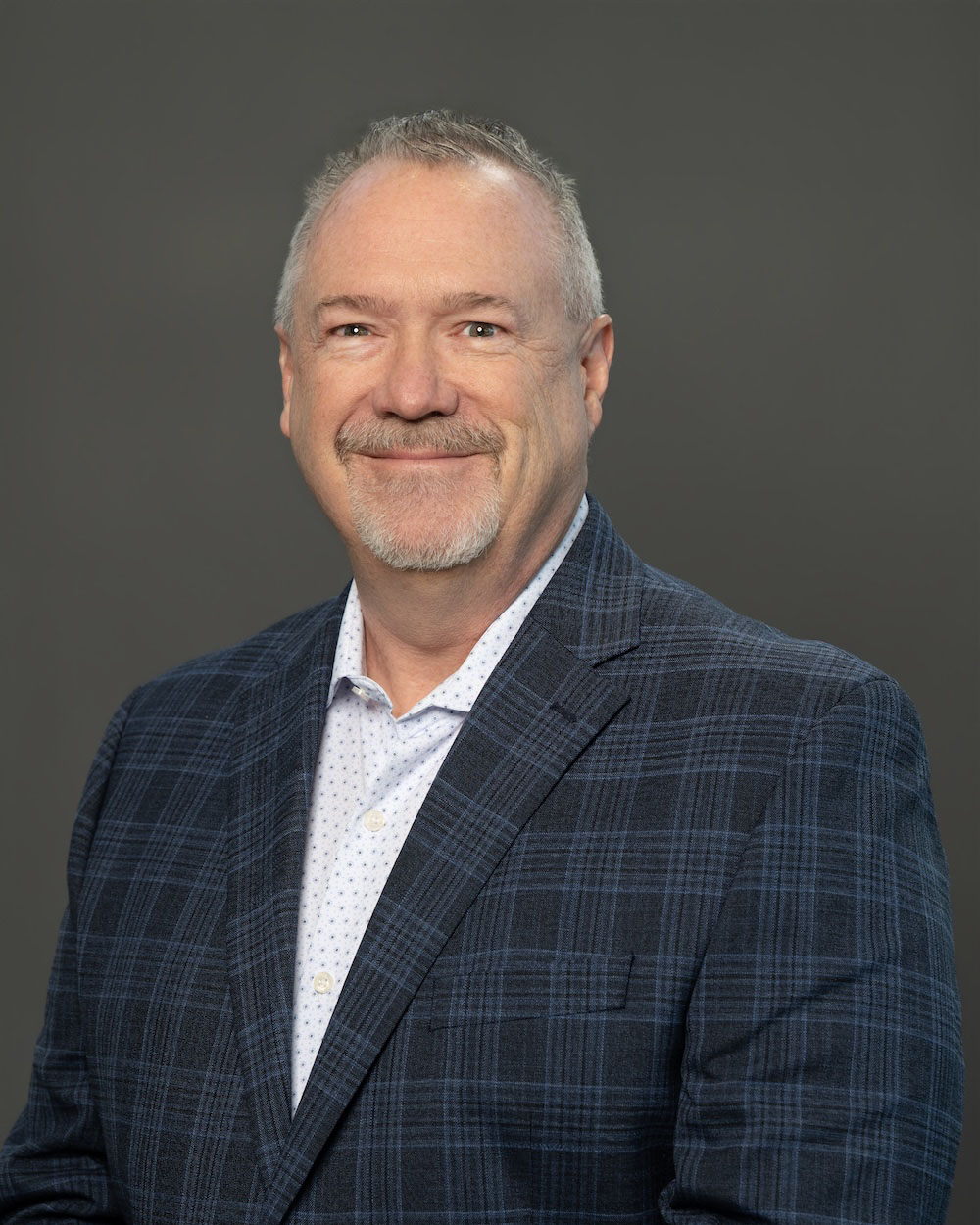 Gene-Burcham-edit Portrait of a smiling middle-aged man with short gray hair and a goatee, wearing a blue plaid blazer and white dotted shirt against a neutral gray backdrop.