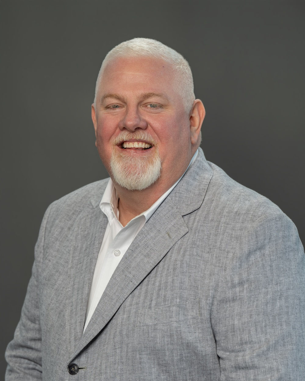 Jason-Street-edit-2 Smiling man in a light gray blazer and white shirt, head-and-shoulders portrait against a dark gray backdrop.