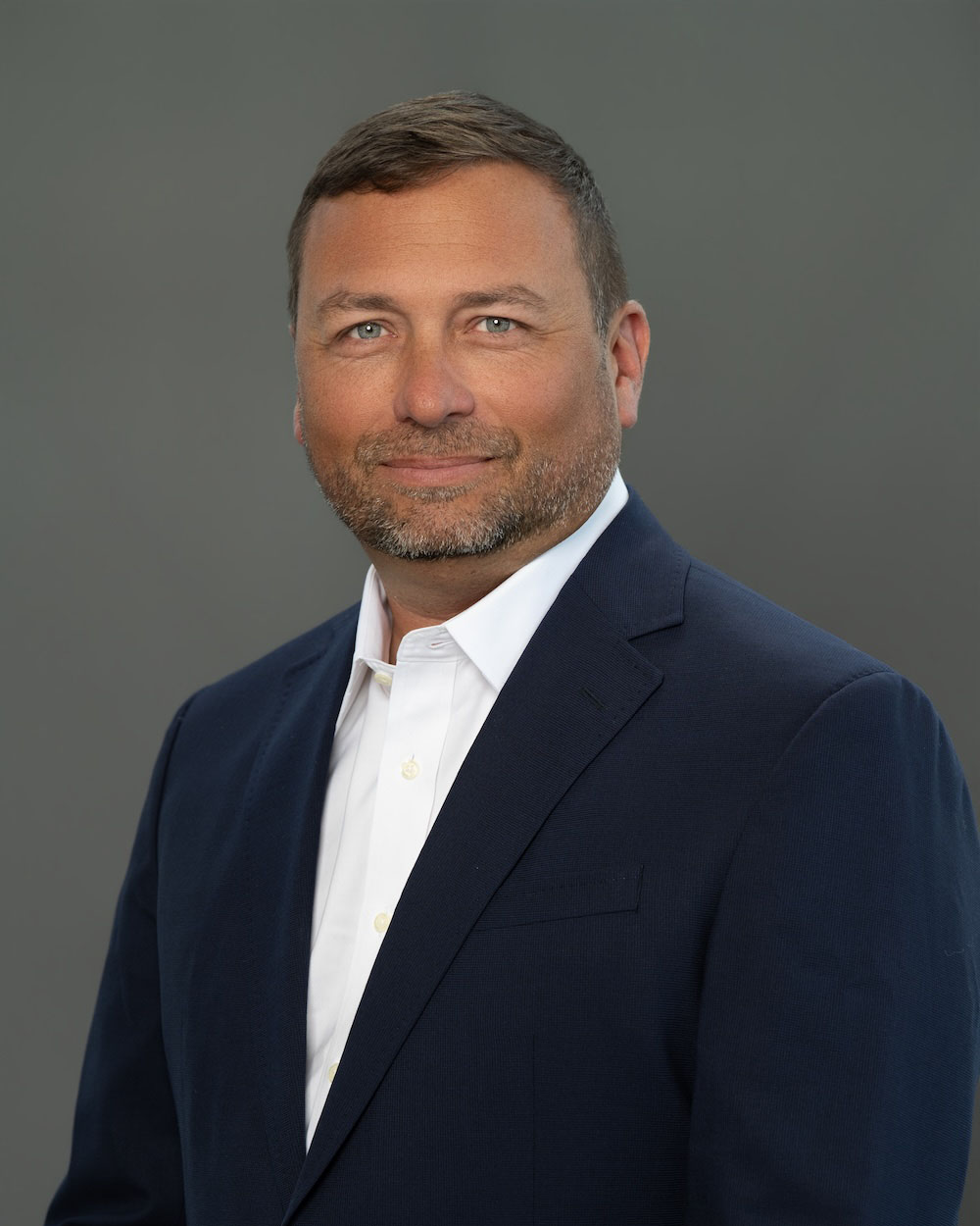 Jerrod-Ehrgott-edit-2 Professional headshot of a man in a navy blazer and white shirt against a gray studio backdrop, smiling slightly.