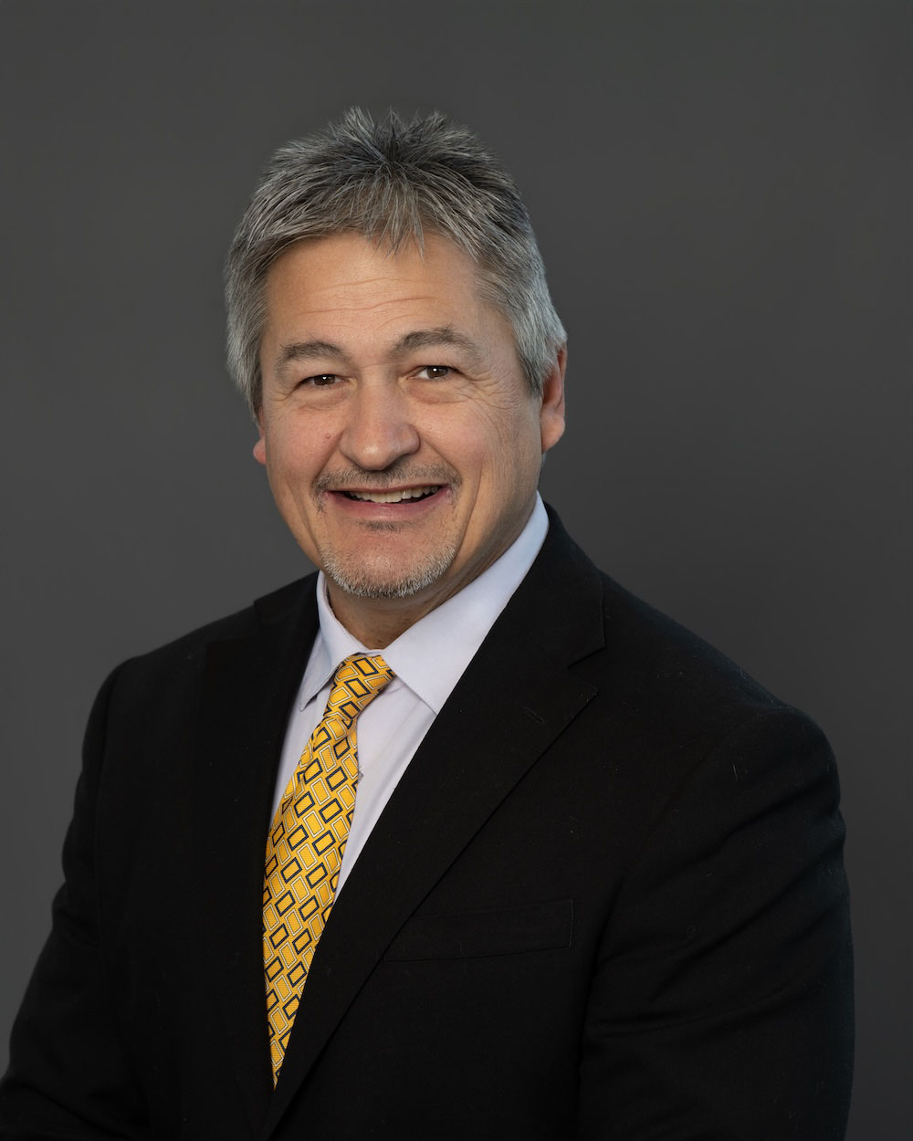 Jon-Acton-edit-2 Professional headshot of a smiling man in a black suit and yellow patterned tie against a gray backdrop.