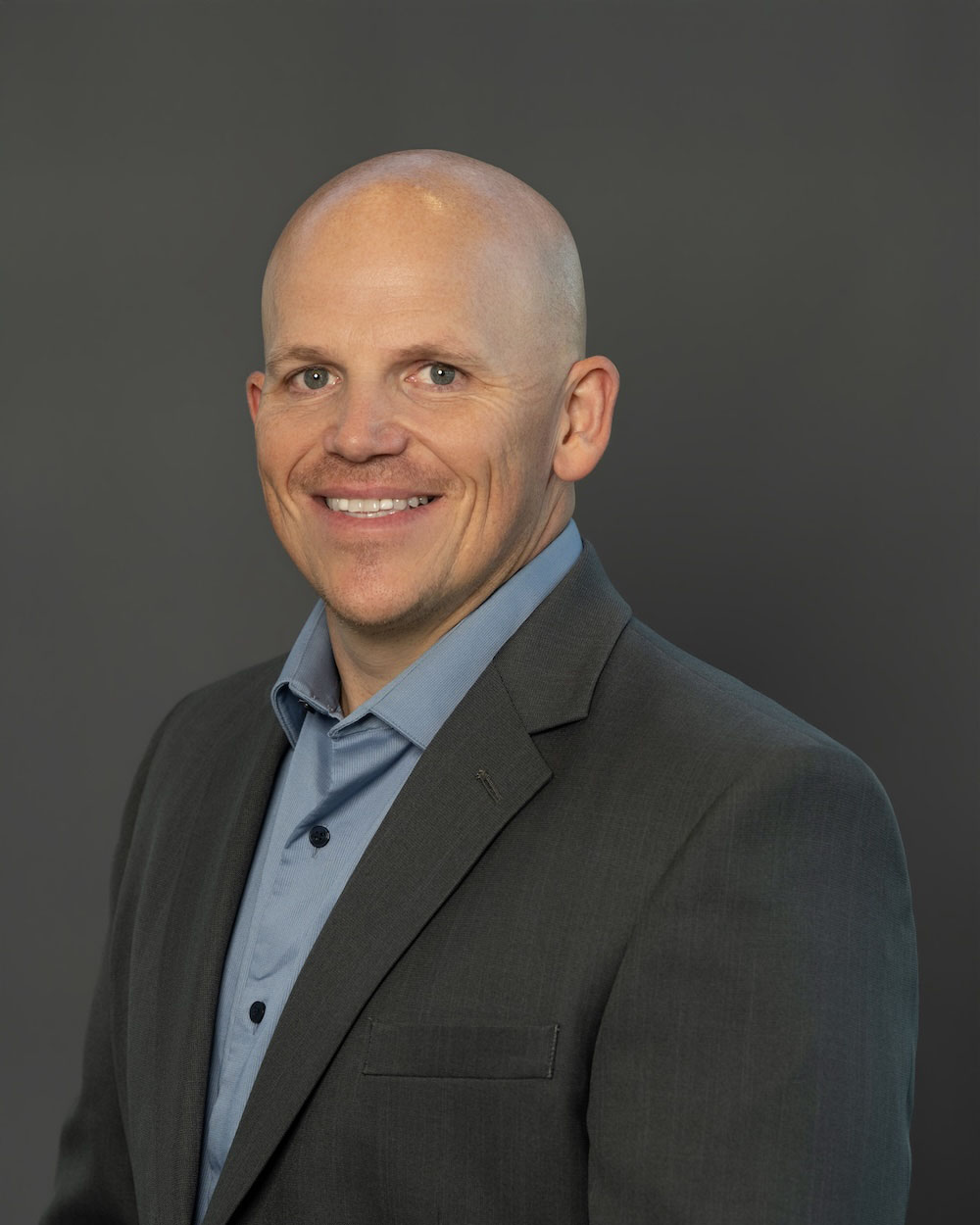 Jon-Murphy-edit-2 Smiling bald man in a dark suit and light blue shirt, posing for a professional headshot against a gray backdrop.