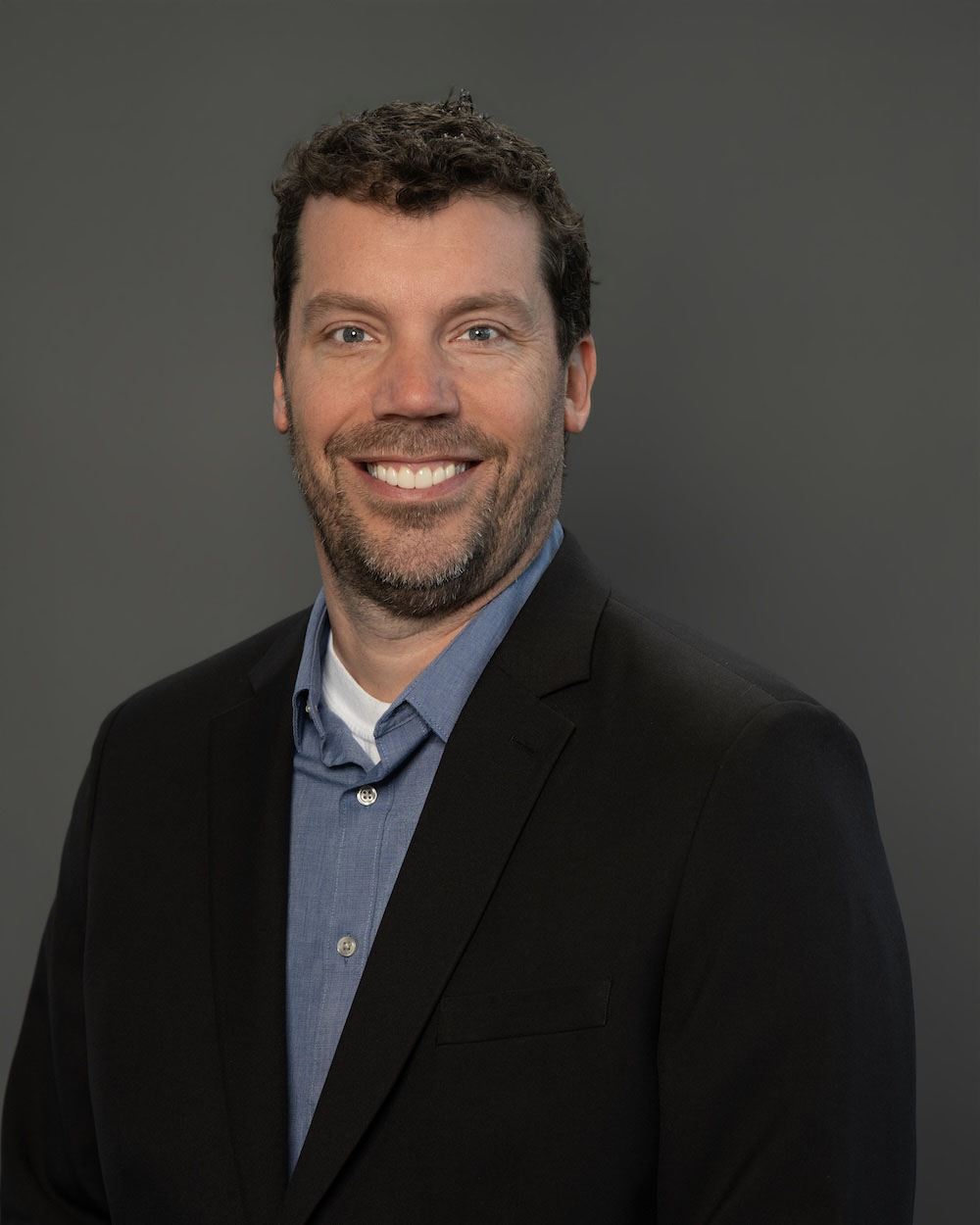 Josh-Leever-edit-2 Smiling man in a black blazer and blue shirt posing for a professional headshot against a gray backdrop.