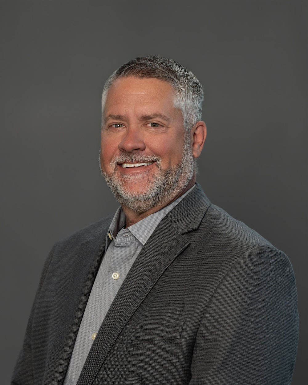 Matt-Hamilton-edit Head-and-shoulders portrait of a smiling man in a gray blazer and light shirt against a dark gray background, a professional headshot.