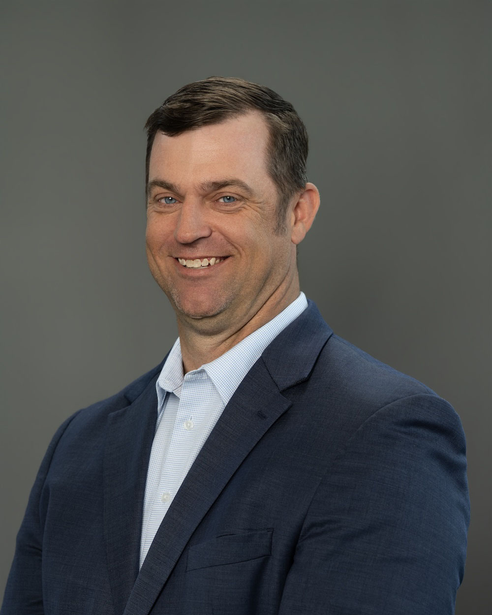 Mike-Mitchell-edit-2 Smiling man in a navy blazer and light blue shirt poses for a professional headshot against a gray backdrop.