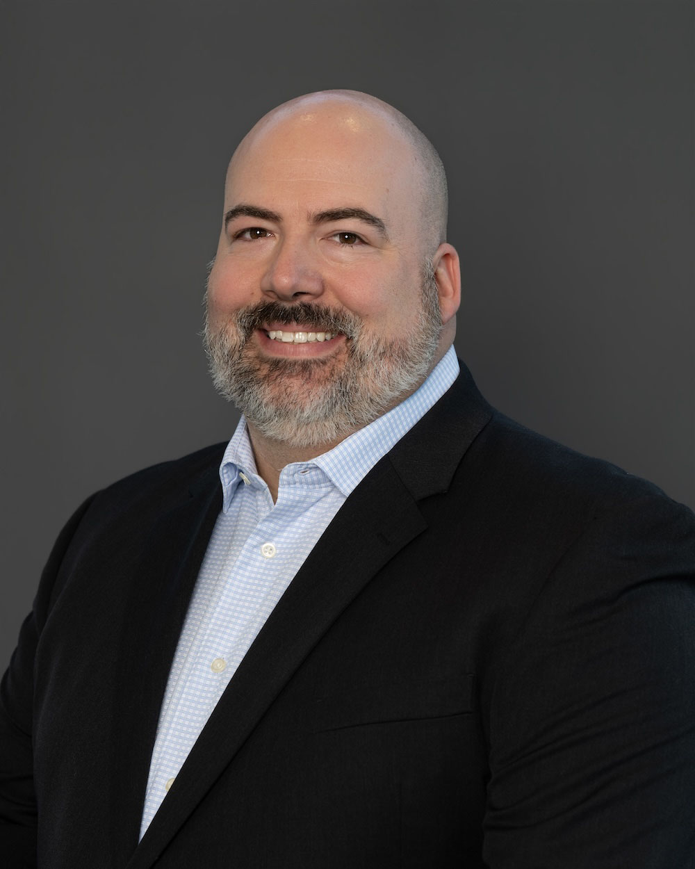 Rick-Saylor-edit-2 Smiling man in a black blazer and light blue checkered shirt against a gray studio background (professional headshot).