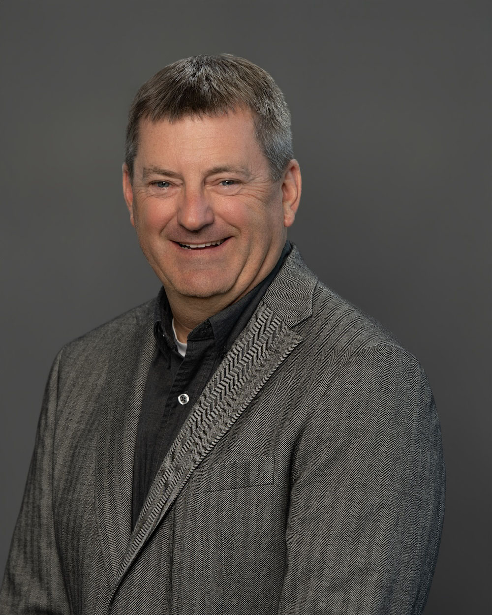 Scott-Cleaver-edit-2 Head-and-shoulder portrait of a smiling man in a gray herringbone blazer against a dark gray background.