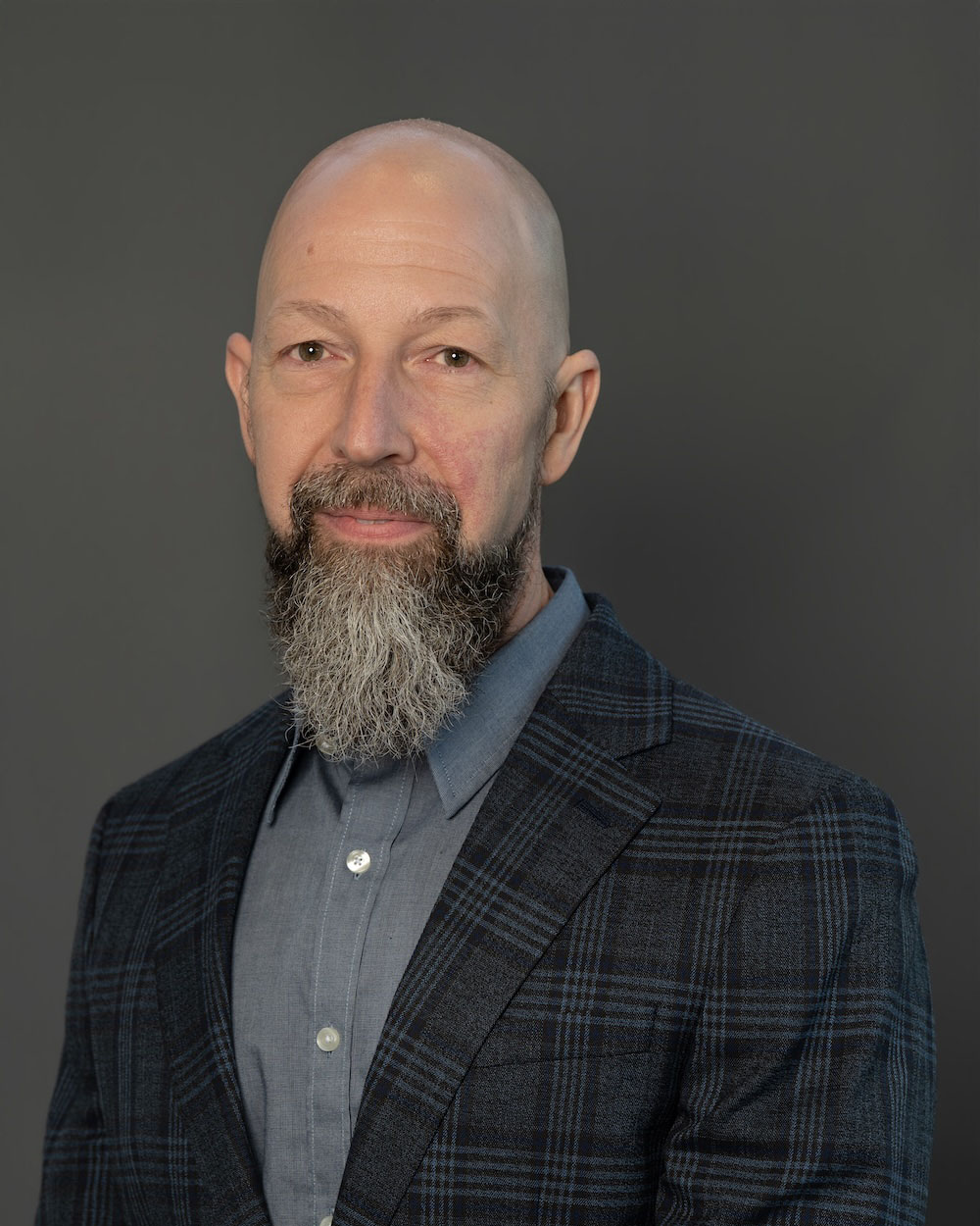 Shane-Foley-edit-2 Head-and-shoulders portrait of a bald man with a gray beard in a blue shirt and navy plaid blazer.