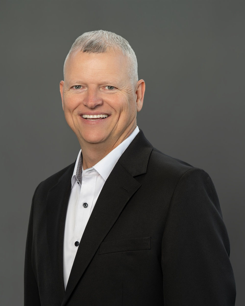 Shane-Pennington-edit-2 Smiling man in a black suit and white shirt poses for a professional headshot against a gray background.