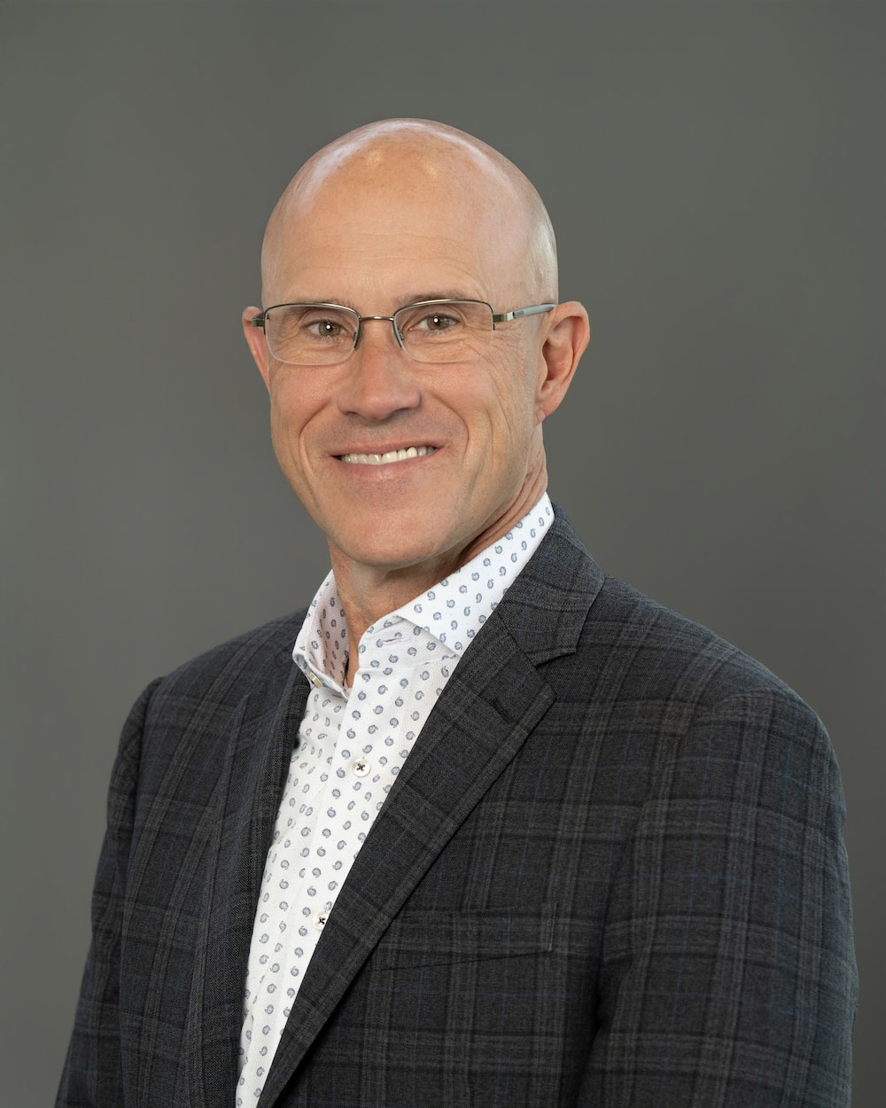Tim-Whicker-edit-2 Professional headshot of a bald man wearing glasses and a dark plaid blazer, smiling at the camera.
