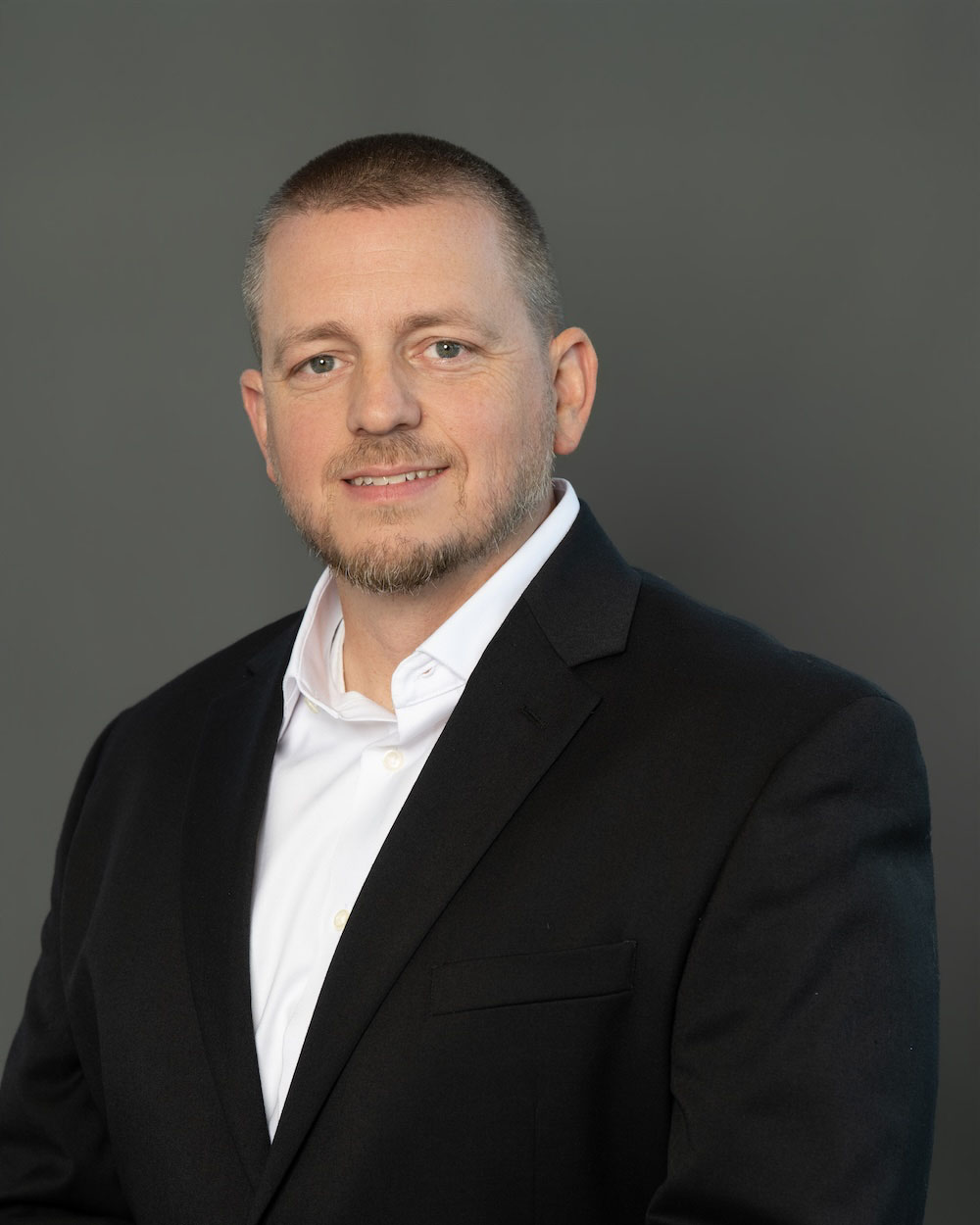 Tom-Johnson-edit-2 Professional head-and-shoulders portrait of a man in a black blazer and white shirt against a gray backdrop.