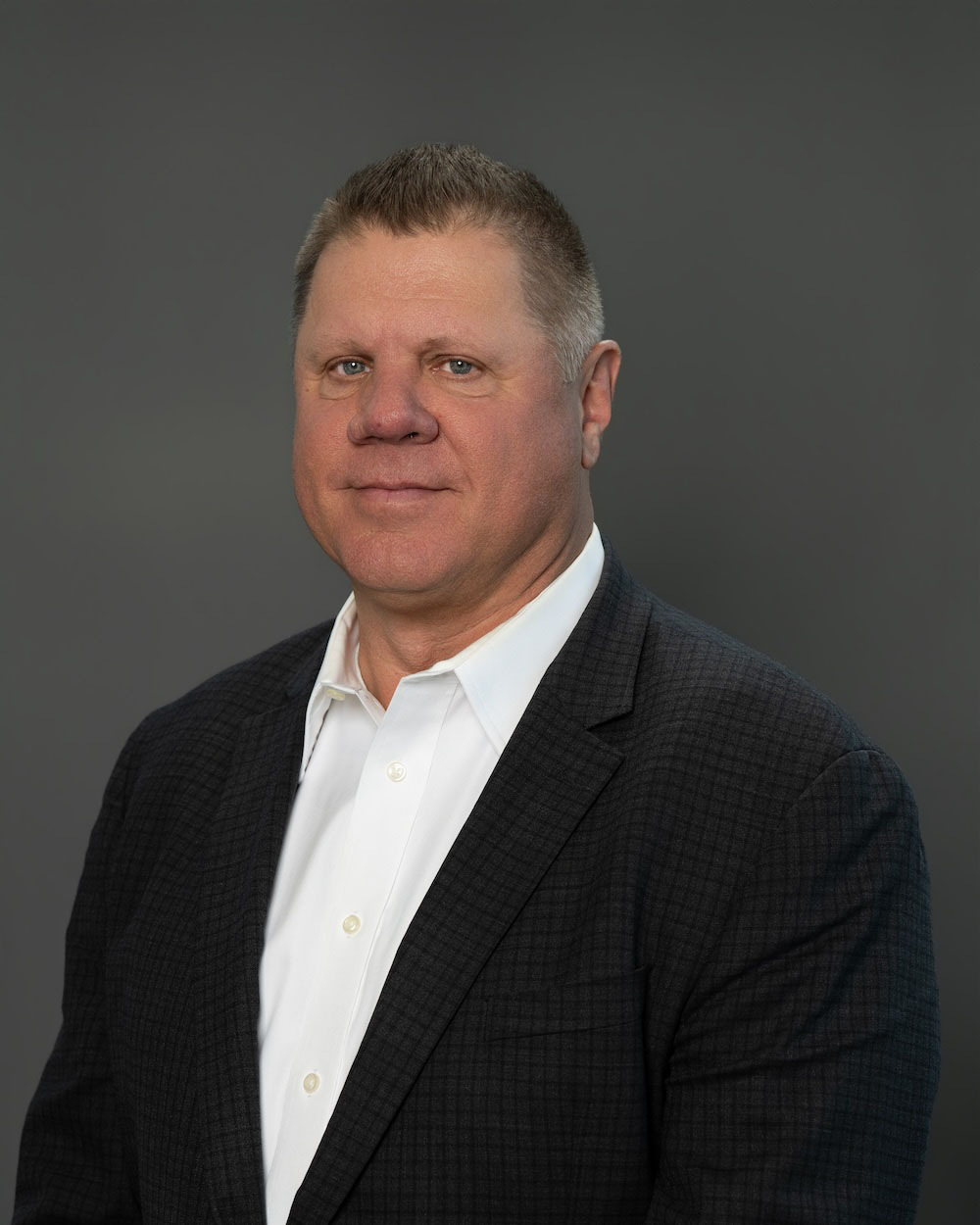 Van-Bragg-edit-2 Professional headshot of a middle-aged man in a dark blazer and white shirt, posed against a gray backdrop.