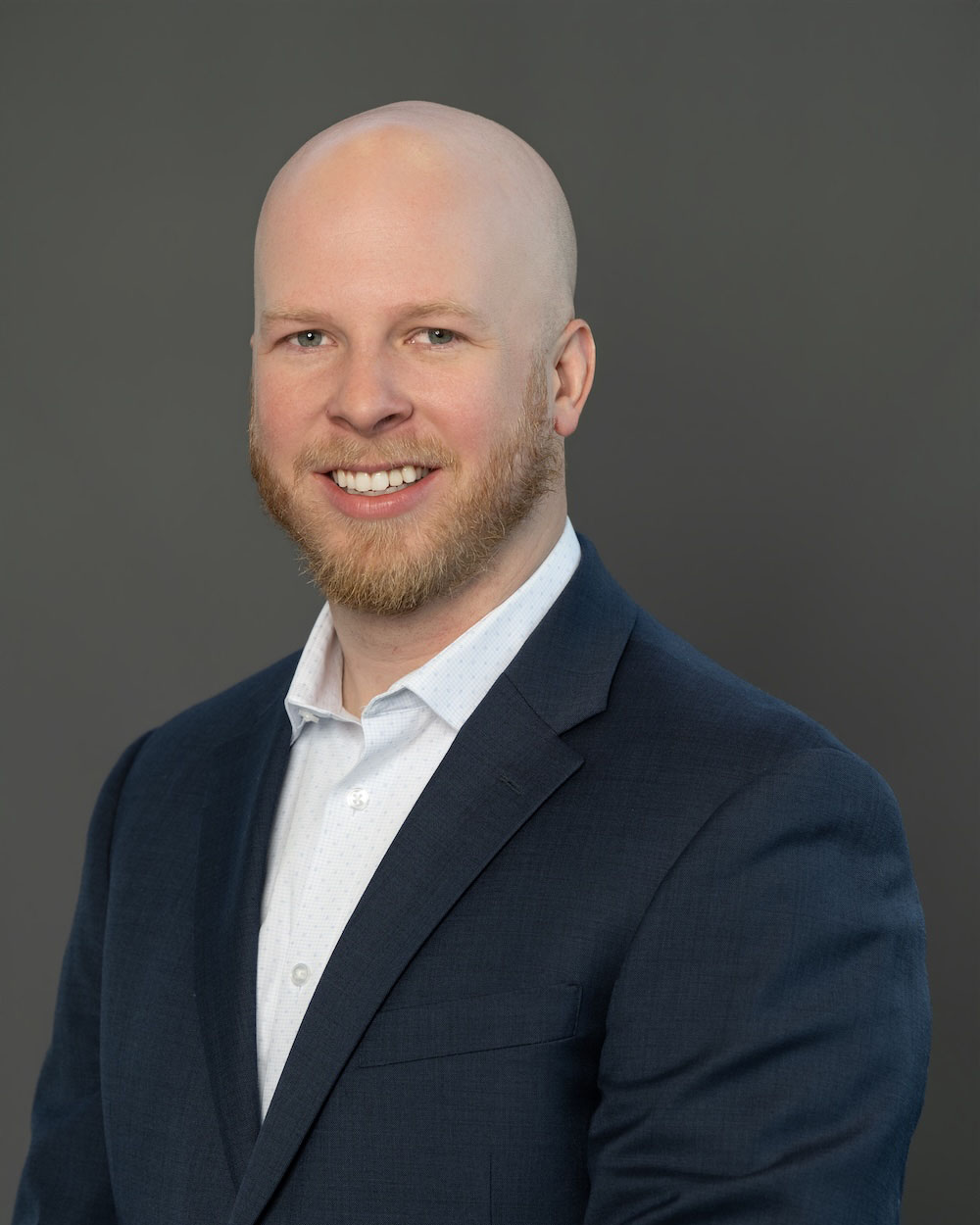 Zach-McCoy-edit-2 Head-and-shoulders portrait of a bald man with a red beard in a navy blazer, smiling against a gray background.